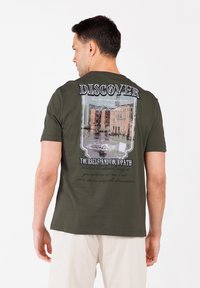 Camiseta de algodón verde oliva con un estampado gráfico de edificios y texto en la parte posterior, de mangas cortas y un corte clásico.