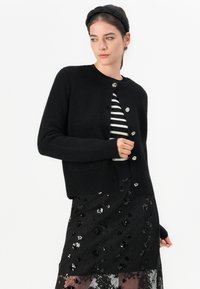 Kvinne som har på seg en svart knappet cardigan over en svart og hvit stripet topp, svart blomstret paljettminiskjørt, og en svart polstret pannebånd.