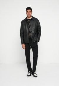 Schwarze Lederjacke mit Knopfleiste, getragen über einem gemusterten Pullover, kombiniert mit schwarzer Hose und zweifarbigen Loafers. Einfacher Hintergrund.