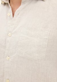 Camicia di lino beige chiaro con colletto a bottoni, taschino singolo sul petto e bottoni chiari. Tessuto strutturato con una finitura sottile e naturale.