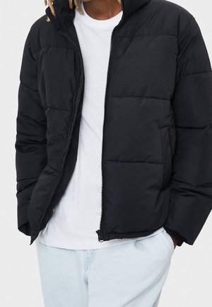 Persoon draagt een zwarte donzen jas over een wit T-shirt met lichtblauwe broek, handen in de jaszakken, tegen een effen achtergrond.