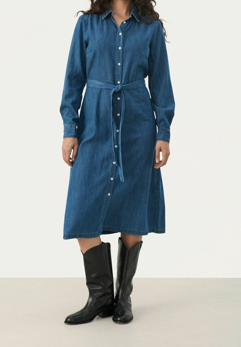 Femme portant une robe en jean à manches longues boutonnée avec une ceinture à la taille et des bottes noires jusqu’aux genoux, debout devant un fond clair et uni.