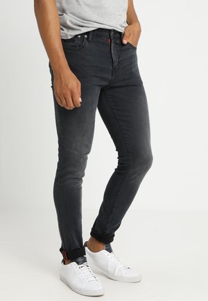 Mężczyzna w czarnych jeansach slim fit z podwiniętymi nogawkami i białych tenisówkach, stoi z jedną ręką w kieszeni na białym tle.