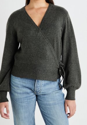 Mujer con un suéter cruzado de color gris oscuro con mangas abullonadas atadas al costado y jeans azul claro, vista desde el cuello hasta las caderas.