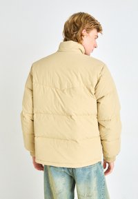 Beige padded jacket with a high collar and elastic cuffs. Features quilted sections for insulation and a relaxed fit, worn over light blue jeans.