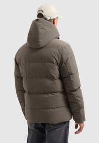 Pufferjack in gedempt bruin, met een grote capuchon, een doorstikt ontwerp en een ontspannen pasvorm, gecombineerd met donkere jeans en een lichte pet.
