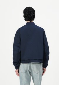 Personne aux cheveux bouclés foncés portant une veste bleu marine et un jean bleu clair, debout de dos sur un fond blanc uni.