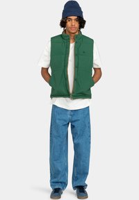 Gilet imbottito verde con colletto alto, indossato sopra una camicia bianca. Abbinato a jeans in denim blu larghi e sneakers scure. Berretto blu in testa.