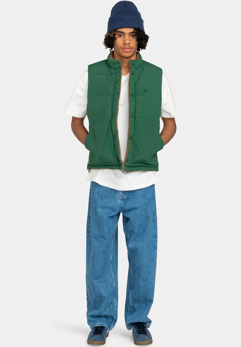 Chaleco acolchado verde con cuello alto, usado sobre una camisa blanca. Combinado con jeans de denim azul holgados y zapatillas oscuras. Gorro azul en la cabeza.