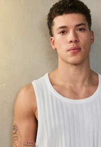 Weißes ärmelloses Shirt mit gerippter Textur, rundem Ausschnitt und ausgeprägten Schulternähten; Tätowierung sichtbar am linken Arm. Glatte Hautfarbe.