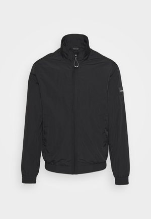 Czarna lekka kurtka zapinana na zamek z wysokim kołnierzem, elastycznymi mankietami i łatką z logo Calvin Klein na lewym rękawie na białym tle.