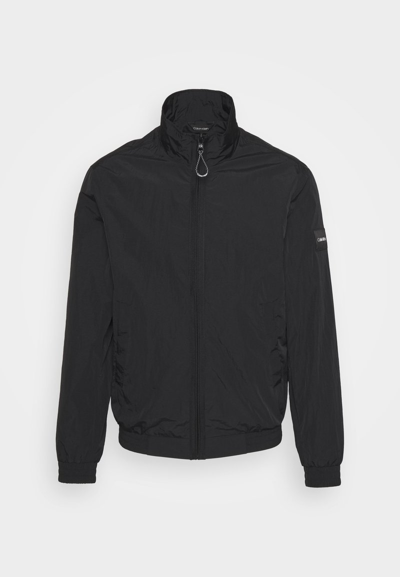 Veste légère noire zippée avec col montant, poignets élastiques et écusson du logo Calvin Klein sur la manche gauche sur fond blanc.