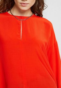 Top in tessuto rosso brillante e liscio con scollatura a chiave. Presenta maniche lunghe e ampie e accenti di cucitura minimi. La collana a catena aggiunge un tocco di dettaglio.