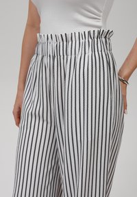 Pantalone s crno-bijelim prugama izrađeni od lagane tkanine, s volanom u struku i bočnim džepovima; vertikalne pruge stvaraju teksturirani izgled.