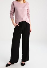 Ljusrosa sweatshirt med texten "ÄG DEN DU ÄR", i kombination med svarta vida byxor. Outfiten har en avslappnad passform och mjukt tyg.