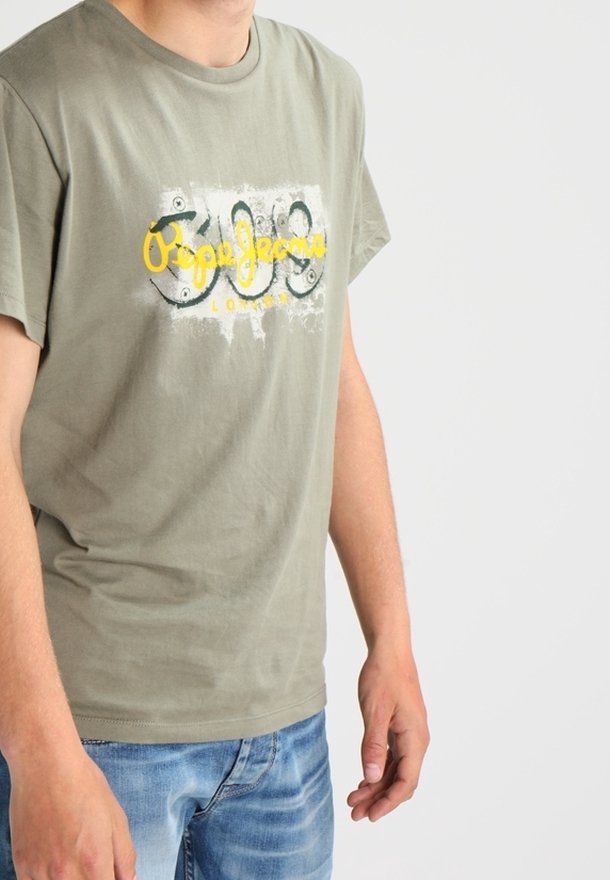 Homme portant un t-shirt graphique vert olive clair et un jean bleu, debout sur un fond blanc uni.