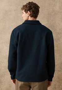 Tmavomodrý vlněný kabát v zmesi s prekladaným golierom, viditeľnými švami, dlhými rukávmi a rovnými okrajmi, nosený s khaki nohavicami.