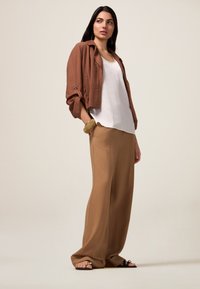 Bruine cropped jack, witte tanktop en beige wijde broek. Sandalen met zwarte banden. Gladde texturen, neutraal kleurenpalet.