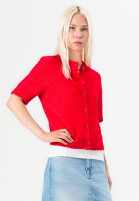 Rode korte mouw cardigan gemaakt van textuur gebreide stof, met een ronde hals en zes knopen aan de voorkant. Gedragen over een wit shirt.