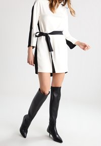 Robe blanche avec des panneaux latéraux noirs et une ceinture. Manches longues et longueur au-dessus du genou. Le modèle porte des bottes à talons aiguilles noires brillantes jusqu'aux genoux.