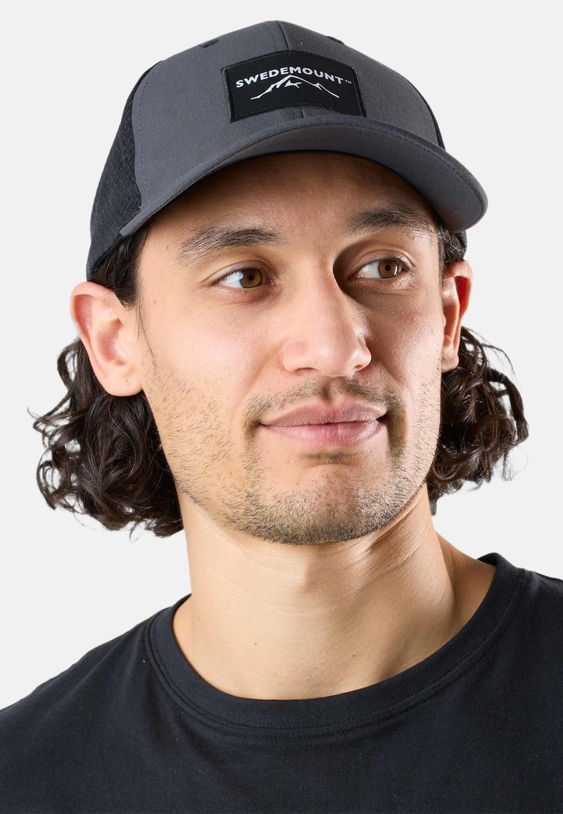 Hombre con barba corta y cabello rizado oscuro que lleva una gorra Swedemount gris y negra y una camiseta negra de cuello redondo, mirando de lado.