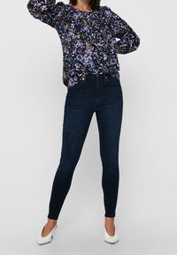 Blusa floreale in tonalità scure con rose viola, abbinata a jeans skinny scuri e tacchi bianchi, caratterizzata da una vestibilità rilassata e polsini elasticizzati.