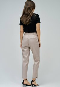 Mujer con cabello largo y castaño de pie de espaldas, vistiendo una camiseta negra de manga corta, pantalones de cintura alta beige y sandalias negras de tacón.