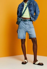 Jeansjacka, grön kort topp, ljusblå shorts, svarta sandaler med vita remmar, teal överkroppsväska. Bakgrunden är ljus orange.