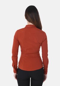 Camicia fitted color ruggine con colletto, maniche lunghe e polsini a bottone. Tessuto liscio con cuciture su misura e orlo curvo.