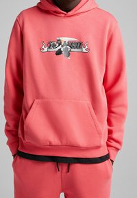 Korallfärgad hoodie med ficka fram, med en grafisk design av en karaktär med ett svärd och fet text. Ribbad manschetter och nederkant.