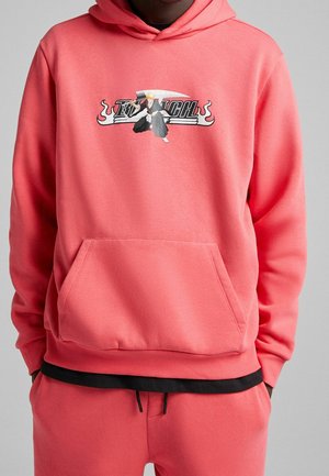 Haut à capuche corail avec poche frontale, présentant un design graphique d'un personnage avec une épée et un texte en gras. Manches et ourlet côtelés.