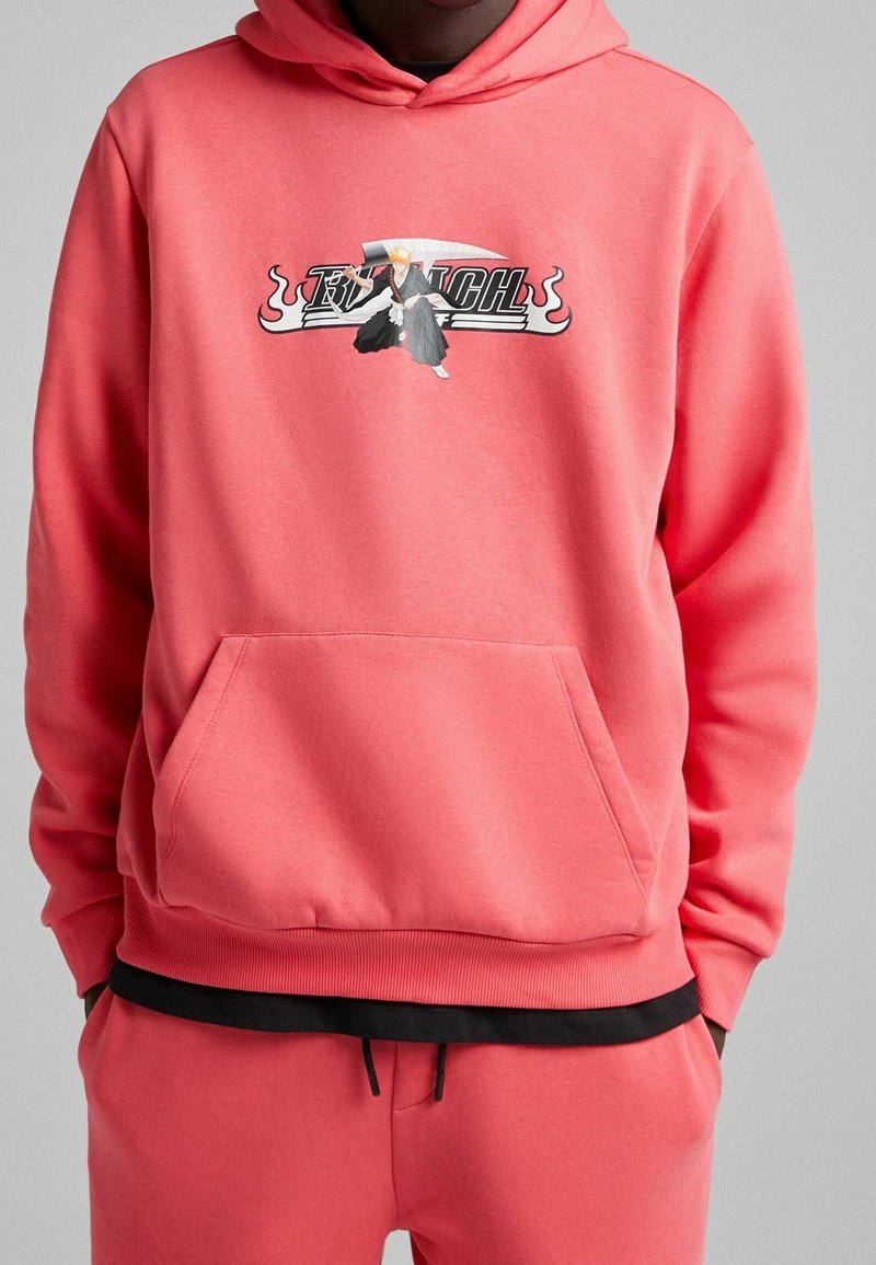Korallfärgad hoodie med ficka fram, med en grafisk design av en karaktär med ett svärd och fet text. Ribbad manschetter och nederkant.