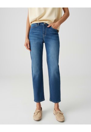Vrouw draagt mid-rise rechte blauwe spijkerbroek met handen in de zakken en beige loafers op een effen lichte achtergrond.