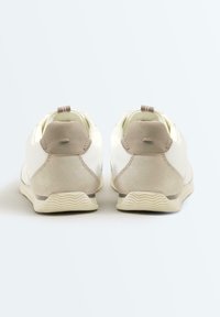Paire de baskets blanches avec des accents en daim beige au talon, vues de dos sur un fond clair uni.