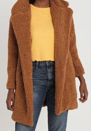 Persoon die een bruine teddyberenjas draagt over een cropped gele trui en blauwe jeans, staand tegen een neutrale achtergrond.