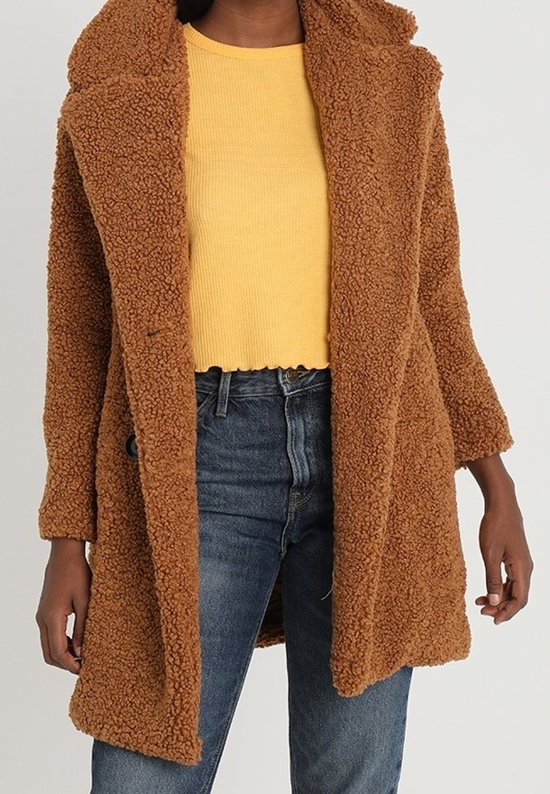 Personne portant un manteau en fausse fourrure marron façon ours en peluche par-dessus un pull court jaune et un jean bleu, debout devant un fond neutre.