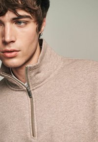Beige Zip-Hoodie mit hohem Kragen, strukturiertem Material und detailliertem Reißverschluss. Verfügt über ein minimalistisches Design mit einer weichen Haptik.