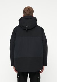 Parka preta com capuz, apresentando um design bicolor, textura de tecido, tiras ajustáveis e bolsos estilo utilitário nas mangas.