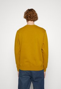 Gul sweatshirt med rund halsringning, ribbade muddar och nederkant. Tyget ser mjukt och bomullsliknande ut. Bärs med blå denimbyxor.