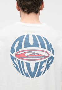 Άτομο που φοράει λευκό πουκάμισο με μεγάλο στρογγυλό λογότυπο Quiksilver σε μπλε και κόκκινο στην πλάτη, από πίσω.