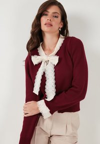 Burgundský pletený svetr s bílým krajkovým límcem a mašlí. Dlouhé rukávy s krajkovým lemováním na manžetách.