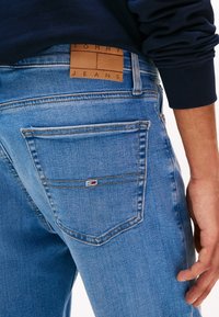 Vue de dos d'une personne portant un jean en denim bleu clair Tommy Jeans avec une étiquette en cuir de la marque au-dessus de la poche arrière, vêtue d'un haut à manches longues bleu marine.