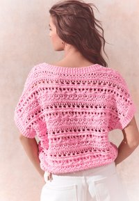 Pink, knitted top with an open, lacy design and short sleeves. Features a wide neckline and a relaxed fit. Worn over white pants.