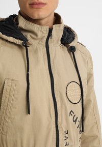 Kurtka w kolorze jasnobrązowym z czarnym kapturem, zamkiem z przodu, troczkami i okrągłą naszywką z napisem "PEACE". Materiał o fakturze z bocznymi kieszeniami.