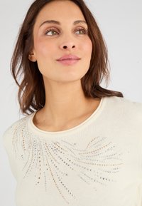 Suéter de color crema con cuello redondo y embellishments decorativos de strass plateados y dorados en un patrón de erupción solar.