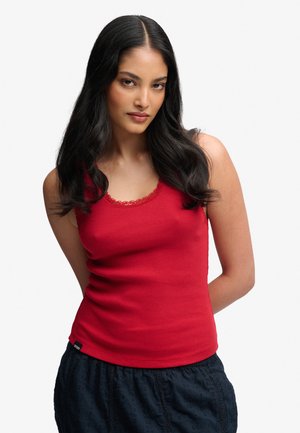 Mujer con cabello largo y oscuro que lleva una blusa roja sin mangas y una falda oscura con textura, de pie con las manos detrás de la espalda sobre un fondo blanco.