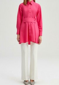 Vestido camisero de manga larga de color rosa brillante con cierre de botones y detalle de cinturón, combinado con pantalones anchos blancos y zapatos gris claro.