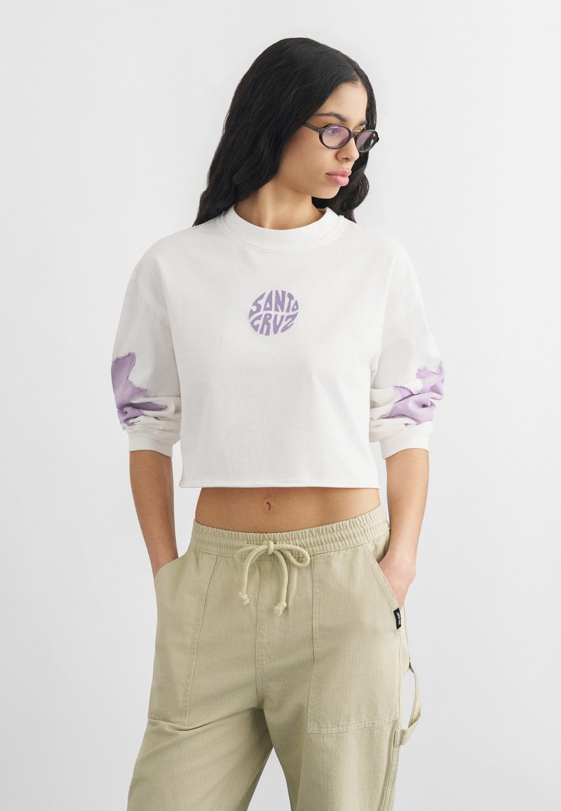 Νεαρή γυναίκα με μακριά μαύρα μαλλιά φοράει στρογγυλά γυαλιά, λευκό κοντό φούτερ με μοβ λογότυπο «Santa Cruz» και μπεζ παντελόνι με κορδόνι.