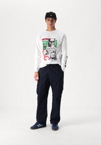 Vit skjorta med lång ärm och grafiska tryck, ihop med marinblå cargobyxor med fickor. Mörkblå sneakers med tre ränder. Svart keps.