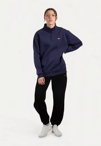 Granatowa bluza na zamek z wysokim kołnierzem, luźnym krojem i prążkowanymi mankietami, zestawiona z czarnymi spodniami dresowymi i białymi trampkami.